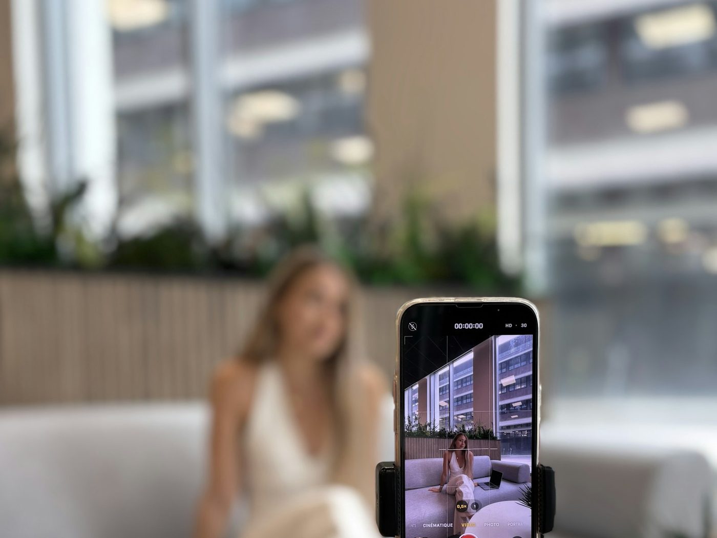 A phone mounted on a tripod recording video, with a person seated on a couch in the background.